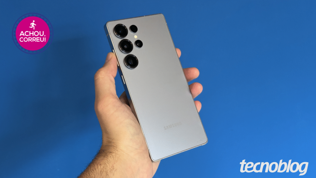 Uma mão segura um smartphone Samsung de cor prateada contra um fundo azul. O aparelho possui um design premium com cantos retos e uma configuração de câmera traseira composta por quatro lentes dispostas verticalmente no canto superior esquerdo.